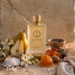 A rectangular glass perfume bottle labeled ZANNIS AFON stands centered on a stone surface, surrounded by pear halves, white flowers, coarse salt, amber honey, a honey dipper, and a textured stone backdrop.