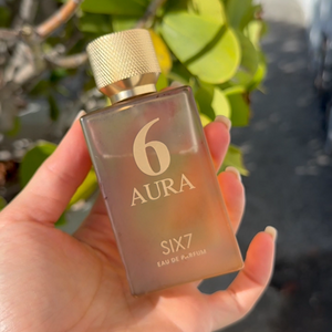 A hand holding a rectangular glass bottle of Aura 6 by Zannis Eau de Parfum with a textured gold cap, set against a background of green foliage and a blurred outdoor scene.