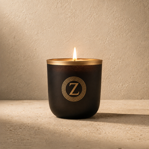 A single lit candle in a dark amber glass jar with a gold rim and a circular gold monogram logo featuring a 'Z' on the front, set against a textured beige wall and surface.