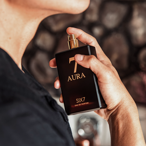 Hand holding a black perfume bottle labeled 'SIX7 Aura' against a blurred background