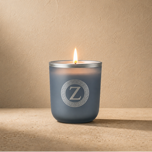 A lit candle in a frosted dark blue-gray glass jar with a silver metal rim and a white circular monogram logo featuring the letter Z, set on a textured beige surface against a matching beige wall.