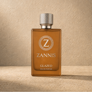 A rectangular amber glass perfume bottle with a textured silver cap, standing on a stone surface against a textured beige wall. The bottle features a silver Greek key and 'Z' logo with the brand name 'ZANNIS' and 'GLAZED EAU DE PARFUM' printed on the front.