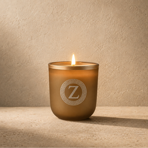 A lit candle in a frosted amber glass jar with a white monogram logo featuring the letter 'Z' inside a Greek key border, set against a textured beige wall and surface.