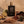 A bottle of Zannis Axios Extrait de Parfum stands centered on a rustic stone surface with ingredients around it, including a lime, ginger root, amber resin, cloves, and dried herbs. The bottle is matte black with a textured gold cap and gold lettering.