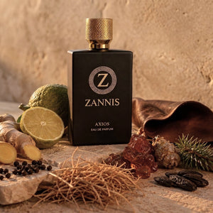 A bottle of Zannis Axios Eau de Parfum stands centered on a rustic stone surface with ingredients around it, including a lime, ginger root, amber resin, cloves, and dried herbs. The bottle is matte black with a textured gold cap and gold lettering.