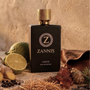 A bottle of Zannis Axios Eau de Parfum is centered, styled with citrus, ginger, amber, and spices on a rustic stone surface against a textured beige background.